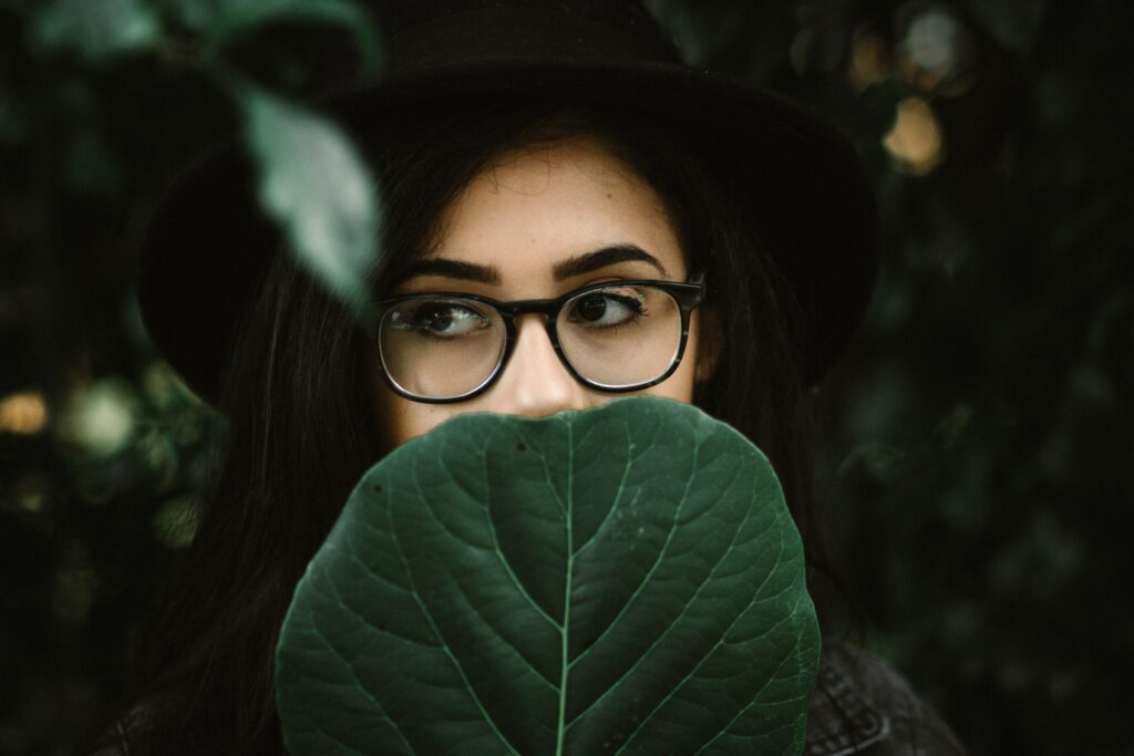 woman with dark hair, black hat, and glasses, holding large dark green leaf over the lower half of her face